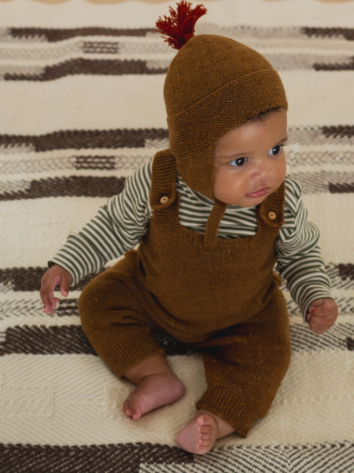 baby in chestnut brown wool salopette with button straps, matching tassel hat and striped top