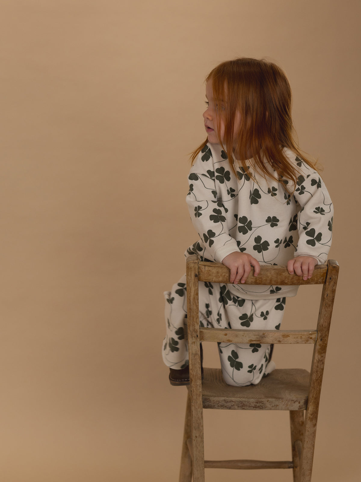 toddler in matching cream sweatshirt and pants with green clover print, kneeling on a wooden chair