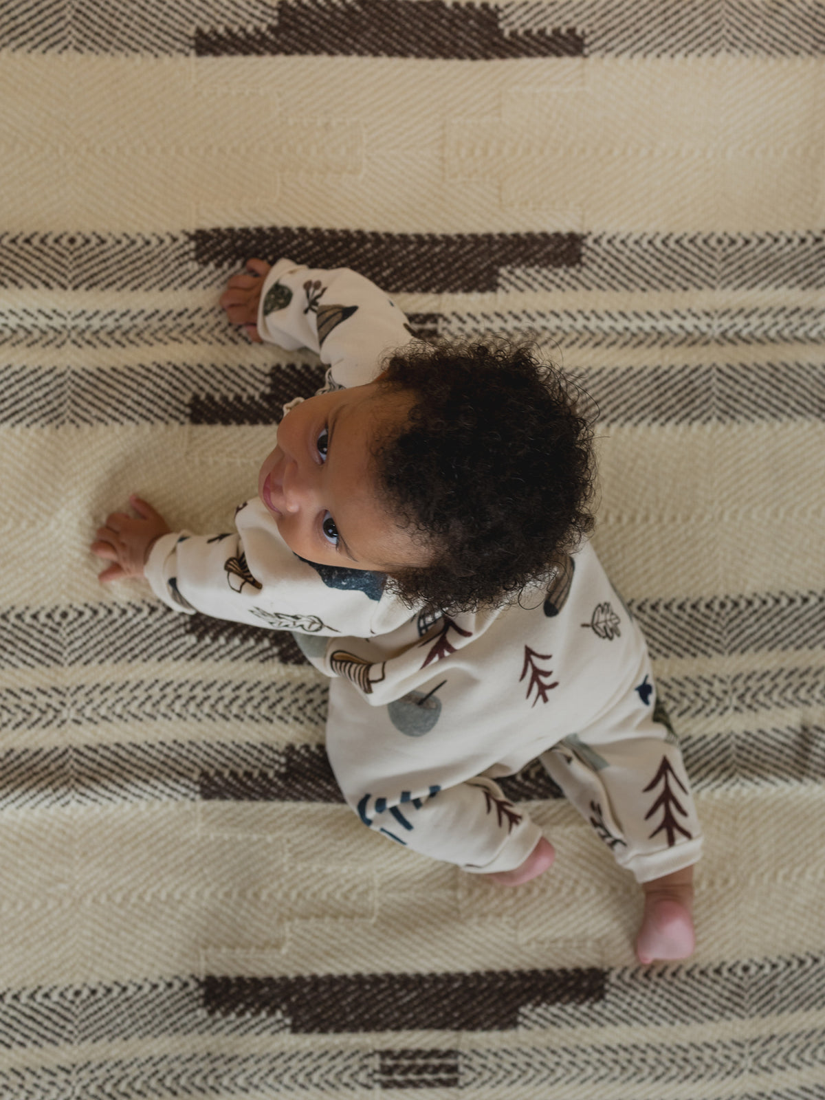 baby in cream undyed cotton onesie with cosy nature-themed hand-drawn prints, crawling on carpet