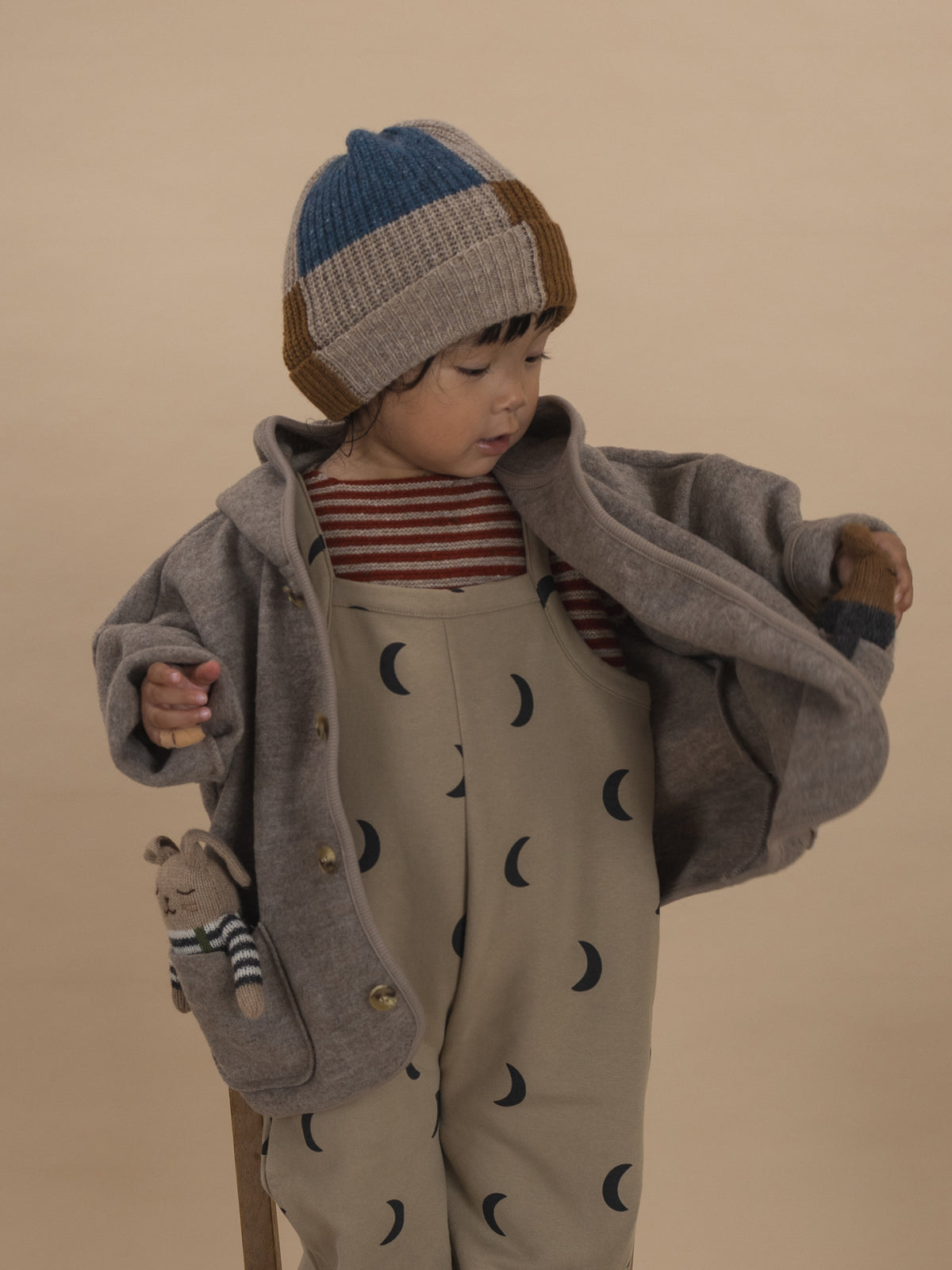 toddler in beige dungarees with black moon print, striped sweatshirt, patchwork beanie, and wool jacket