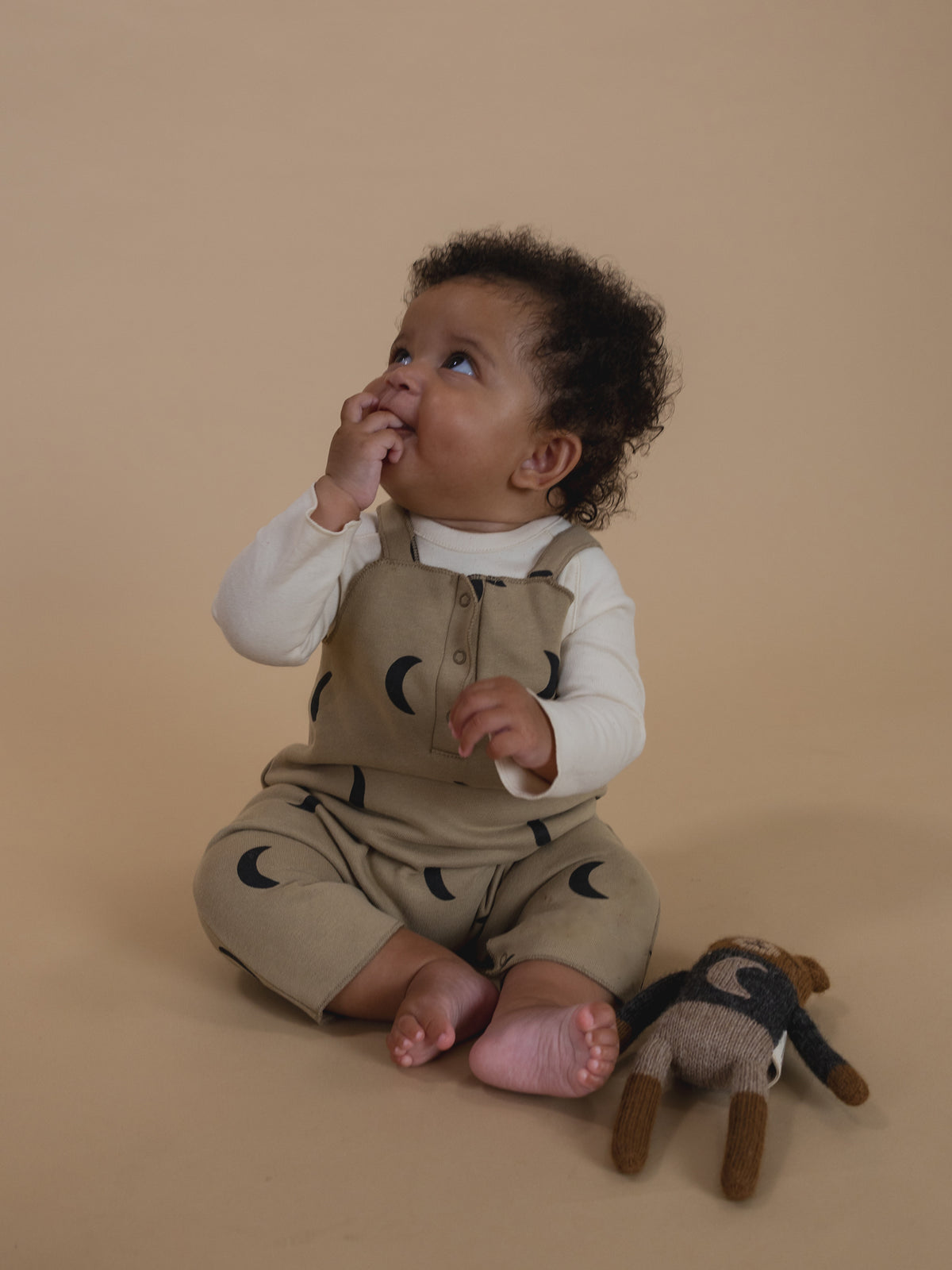 baby in dusky beige salopette with black crescent print and white sweatshirt, sitting with plushie