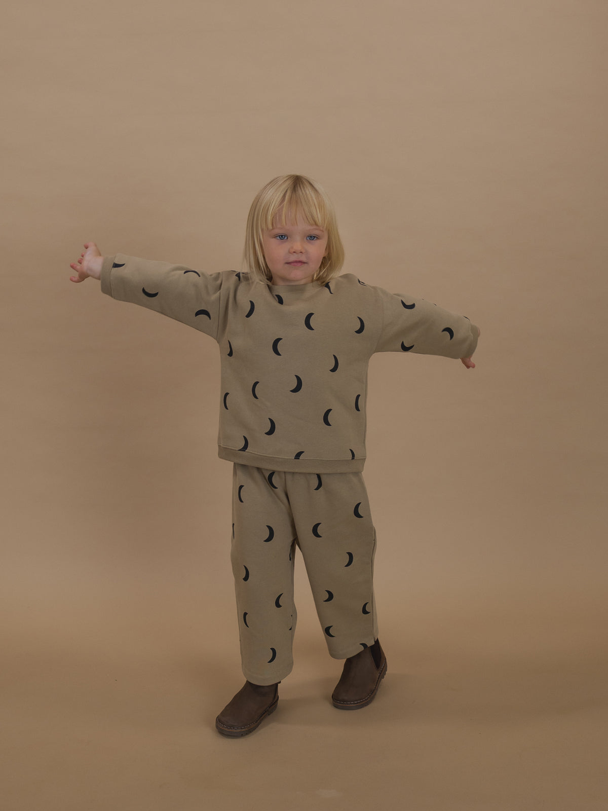 toddler in dusky beige trousers with black crescent print and matching pants, standing with arms out
