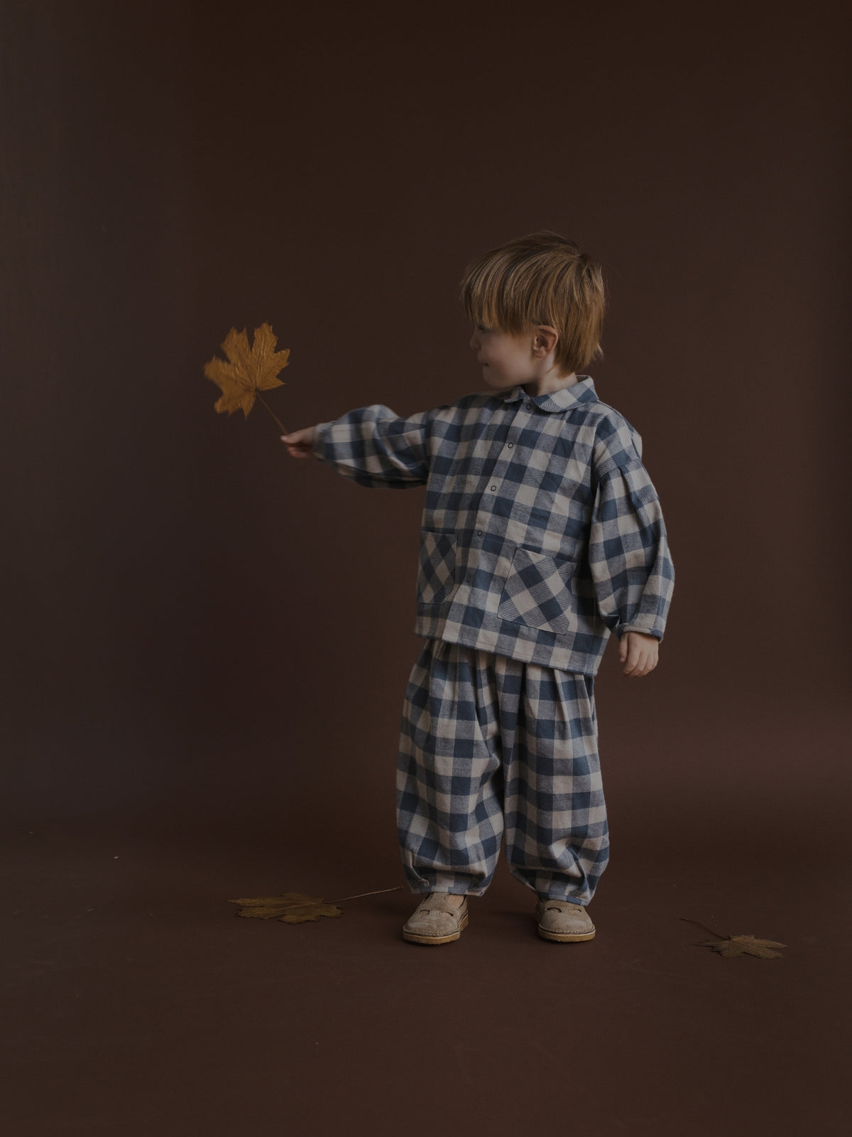 A toddler stands against a dark brown studio backdrop wearing a matching blue and white gingham set, holding a single dry leaf in one hand.
