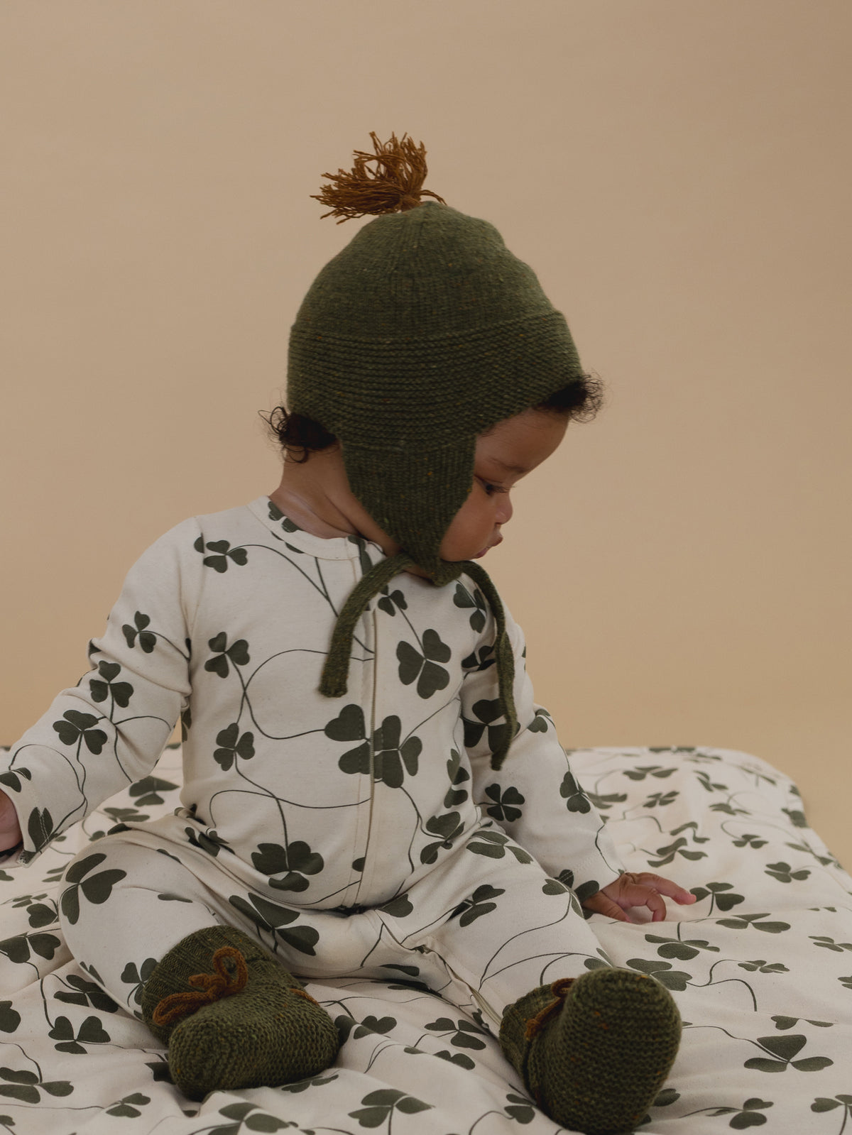 baby wearing forest wool hat with brown tassel and matching booties, and clover sleepsuit looking down