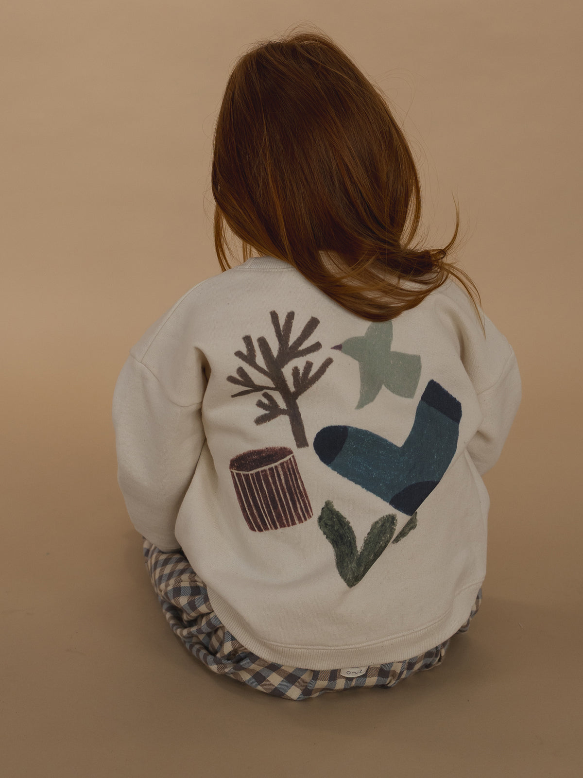 toddler wearing cream cotton sweatshirt with nature-themed back graphic and gingham pants, sitting