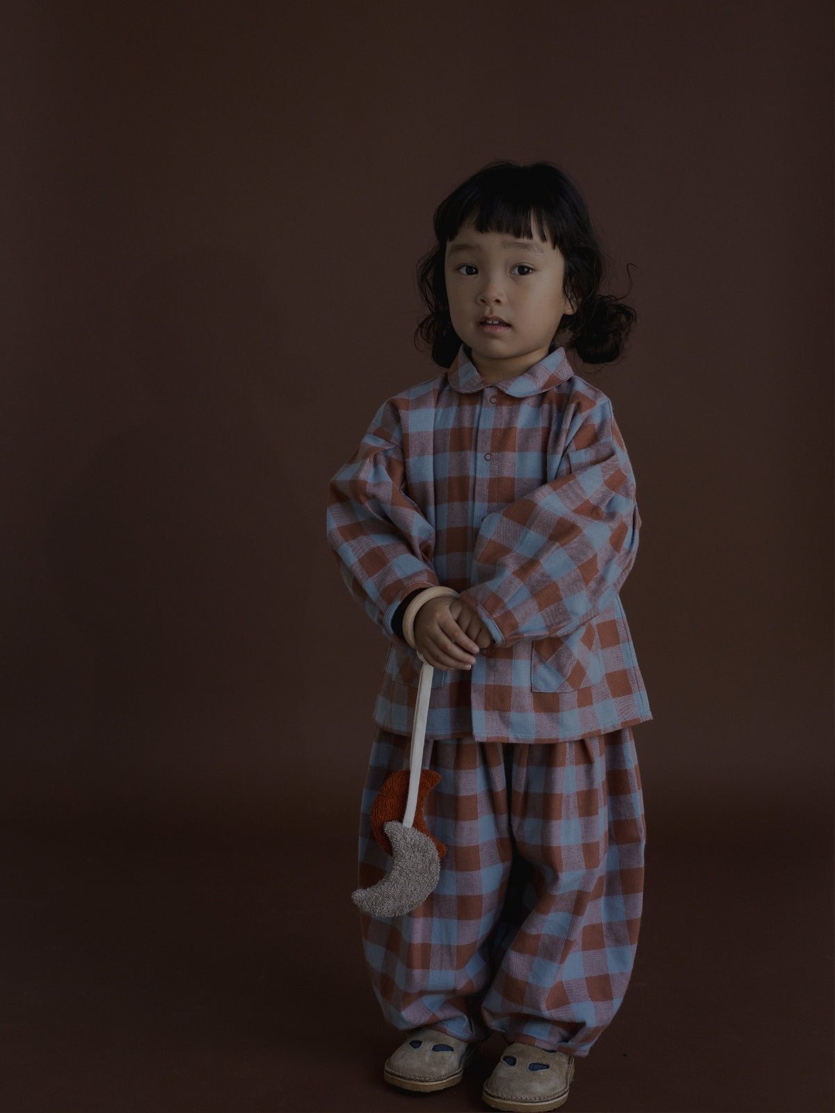 A young child stands against a dark brown studio background wearing a matching plaid pajama set in dusty pink and light blue. The outfit consists of a long-sleeve button-up top and loose pants, and the child is holding a small gray crescent moon toy.