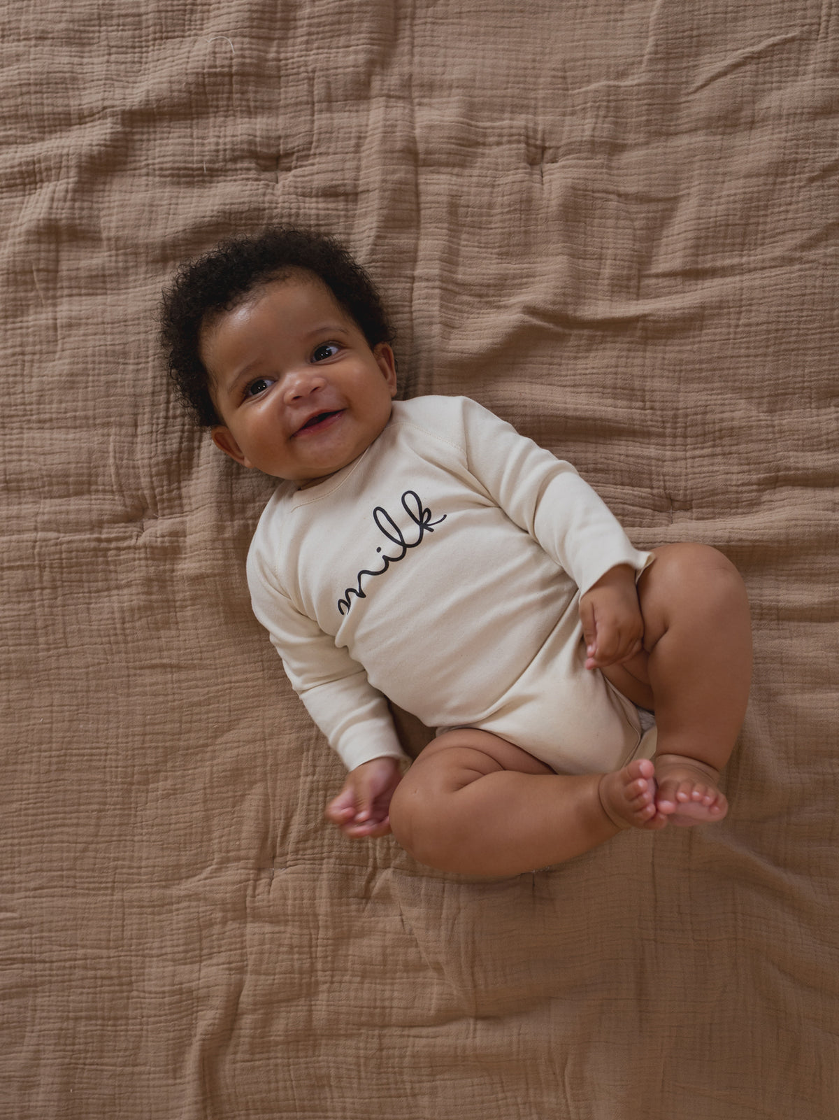 baby wearing cream undyed cotton bodysuit with milk text, lying on muslin blanket