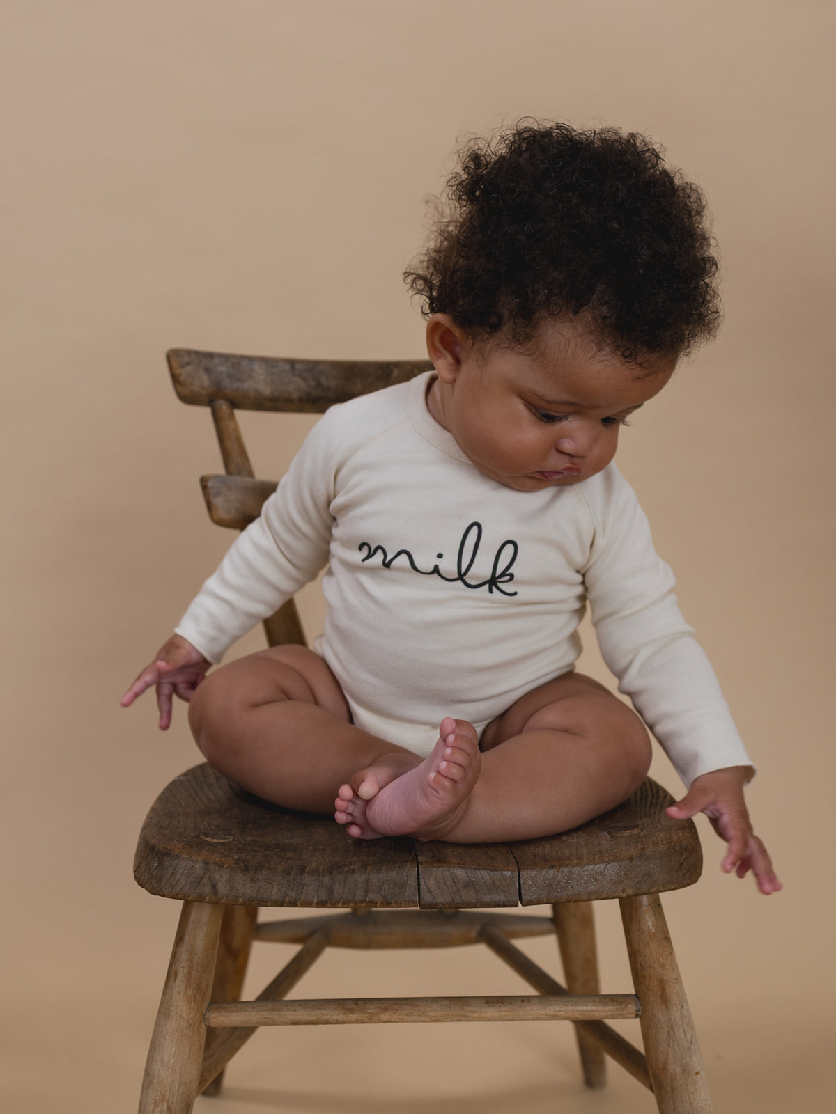baby wearing cream undyed cotton bodysuit with milk text, sitting on a wooden chair