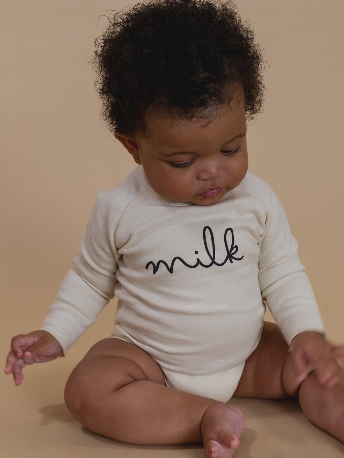 baby wearing cream undyed cotton bodysuit with milk text, sitting on the floor