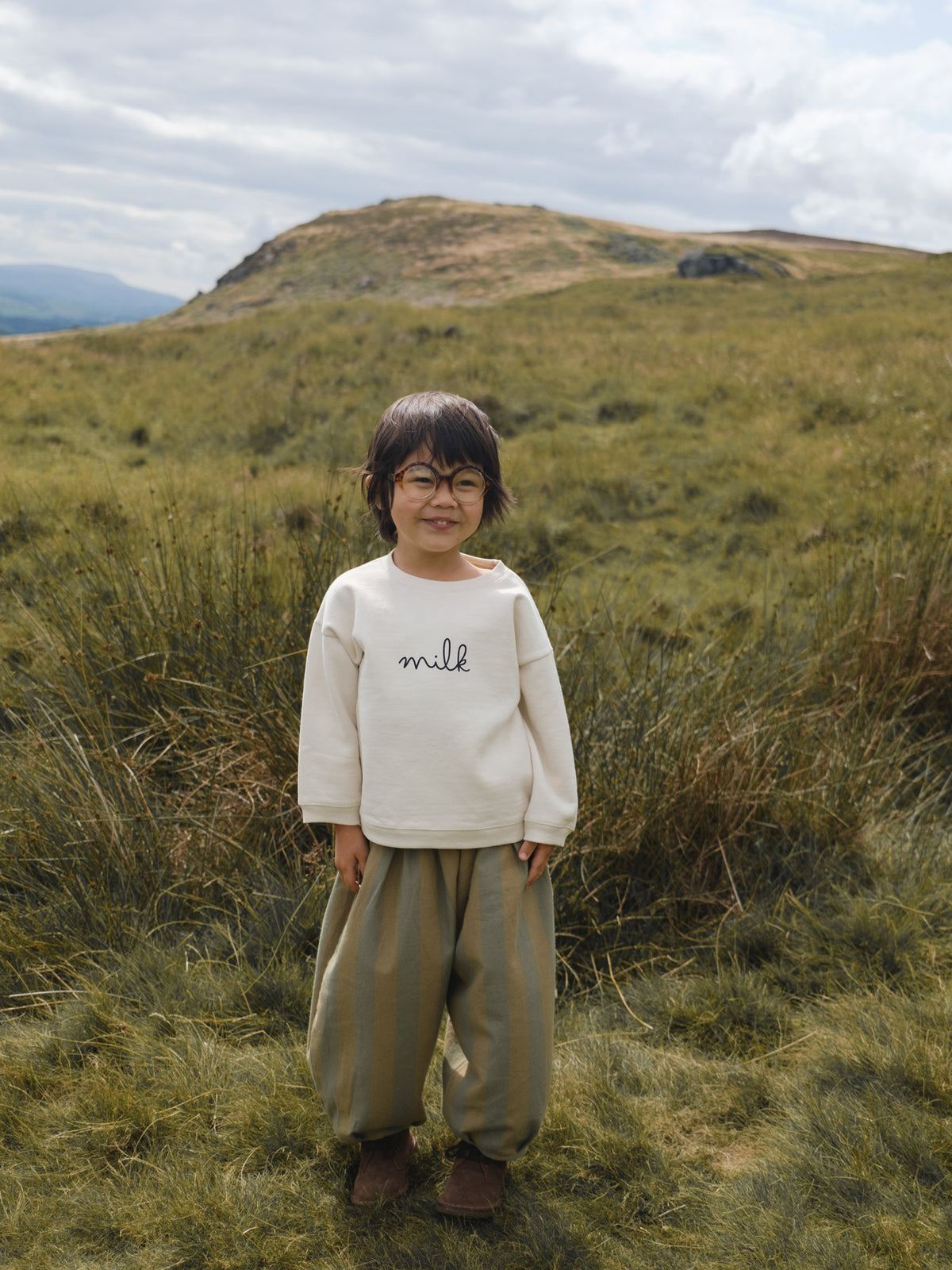toddler in boxy drop shoulder sweatshirt with balloon pants and glasses, in grassy meadow landscape