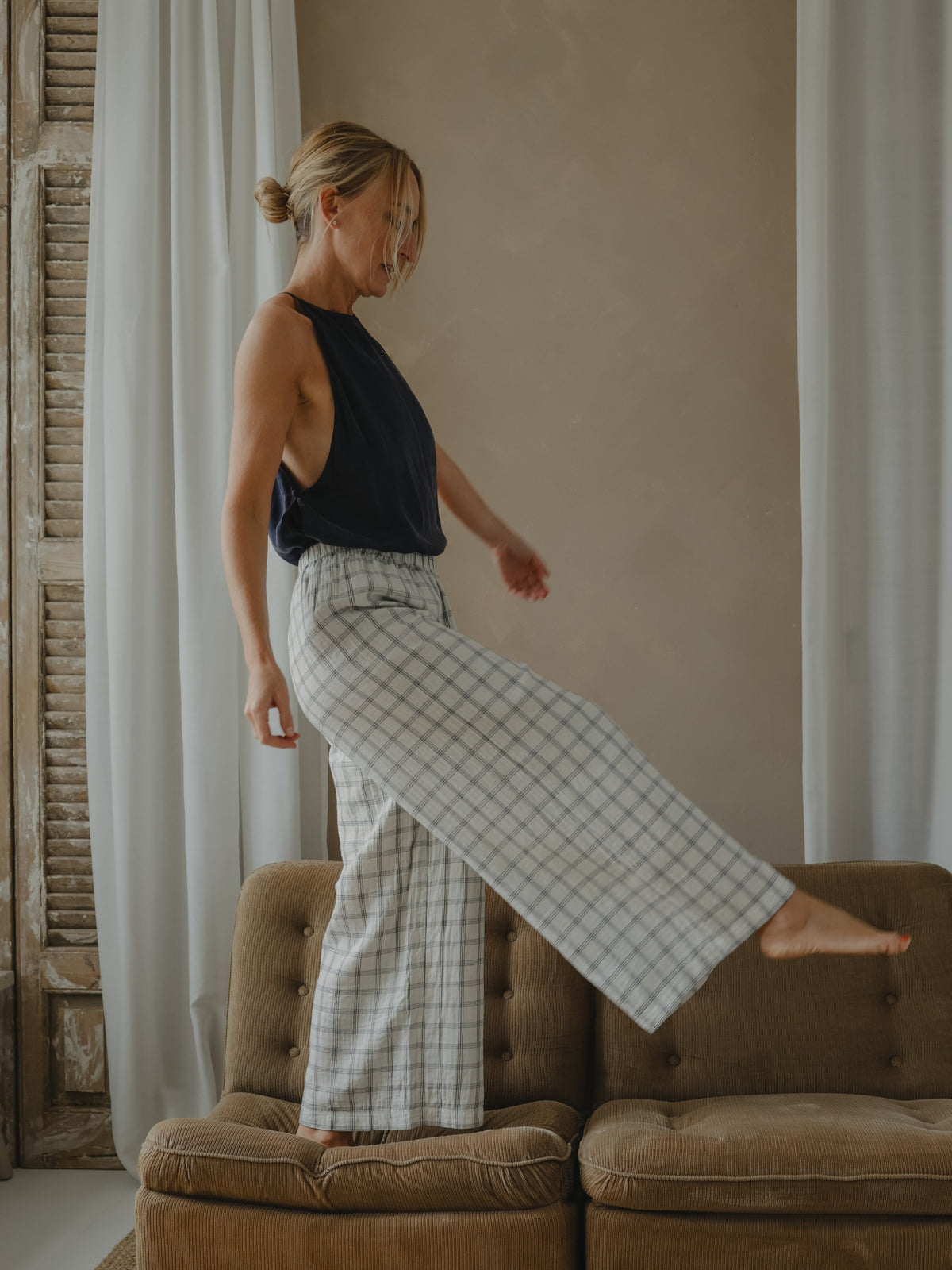 Woman in a black top and checkered pants standing on a brown couch with a neutral wall and curtain in the background.