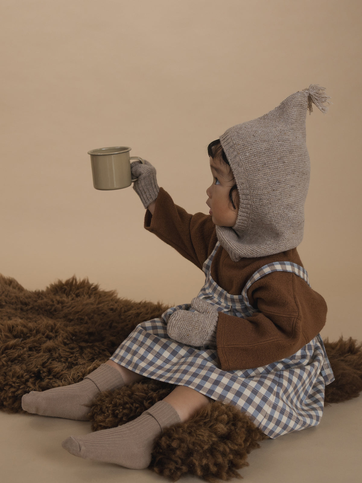 toddler in oatmeal mittens with matching balaclava, brown jumper and gingham skirt, holding cup