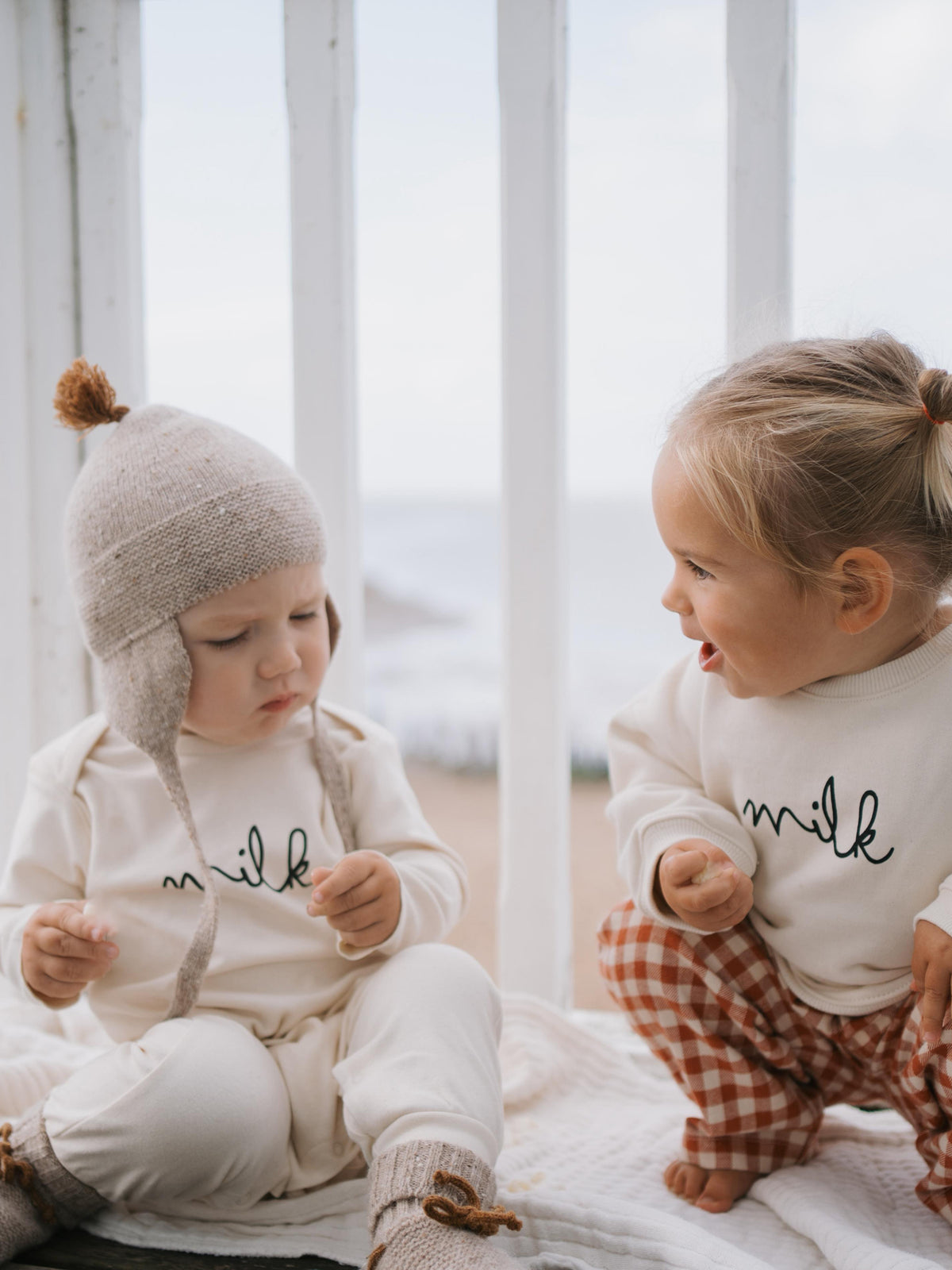 baby in cream playsuit with milk slogan, sitting beside a toddler in cream sweatshirt with milk slogan