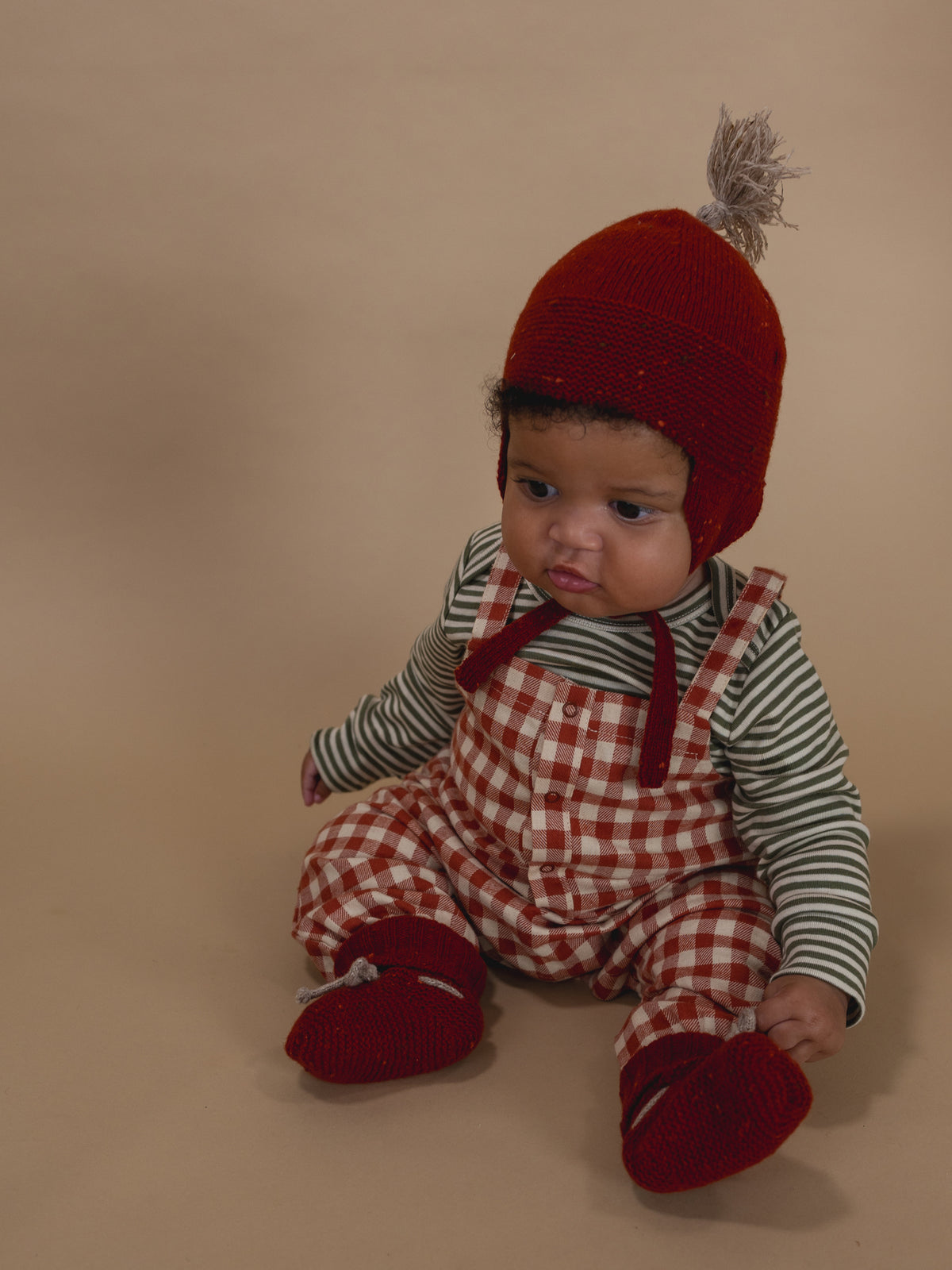 baby wearing red wool booties, matching tassel hat with chin tie, gingham dungarees and striped top