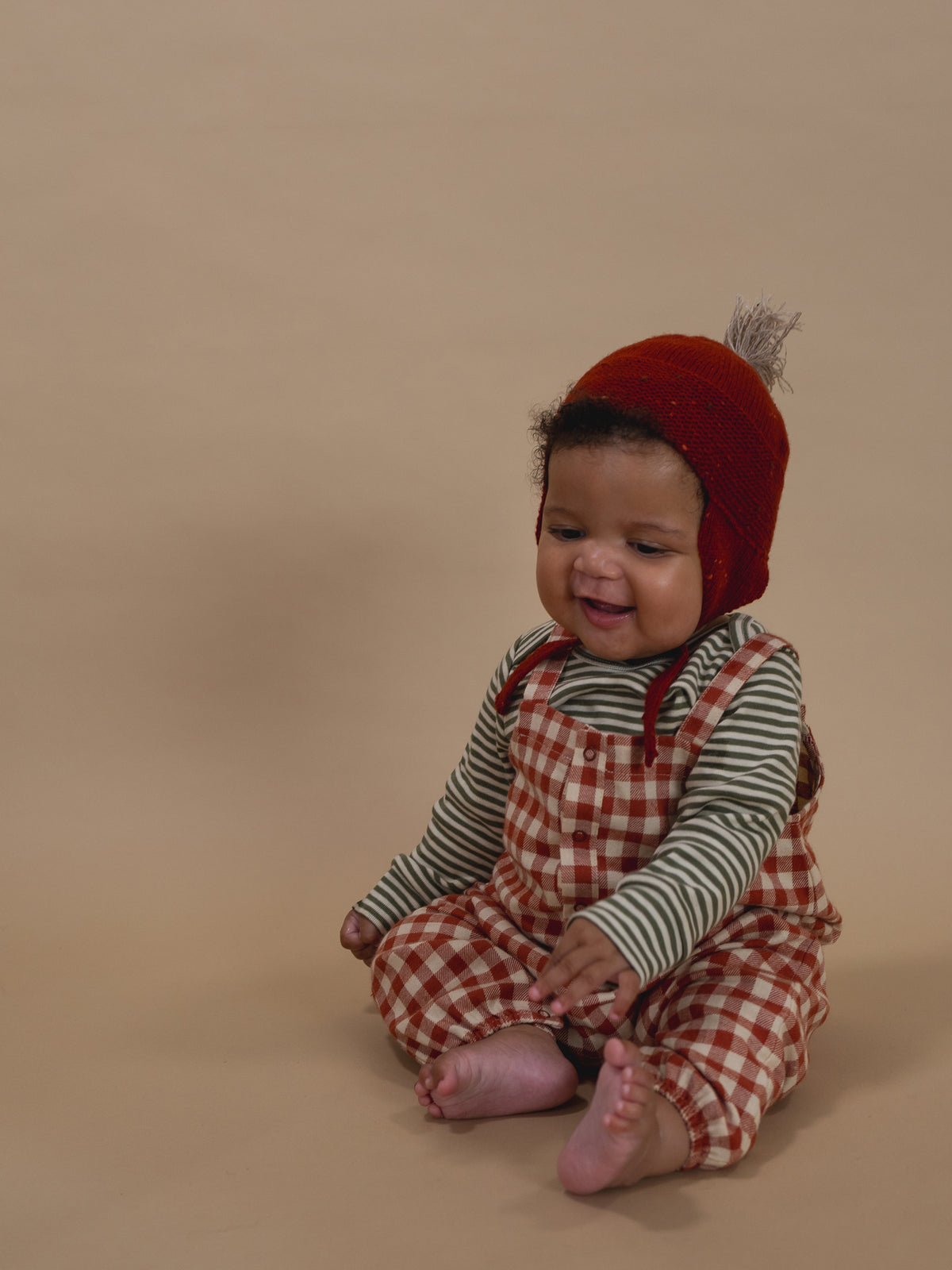 baby smiling in paprika red wool tassel hat, matching booties, gingham dungarees and striped top