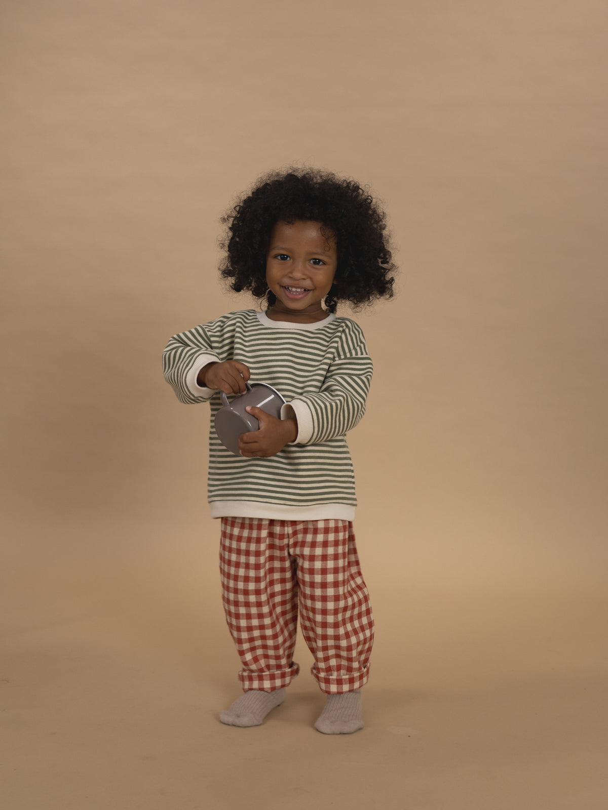 toddler in red and cream gingham carrot pants and green striped sweatshirt holding cup