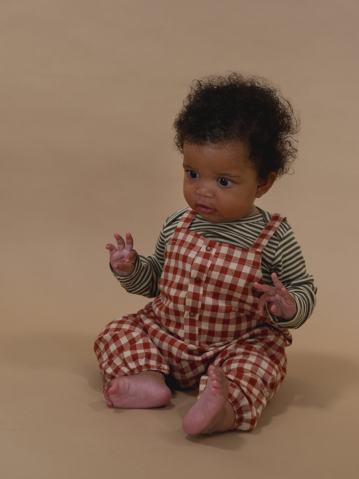 baby in paprika red and cream gingham salopette with green striped sweatshirt, sitting on floor