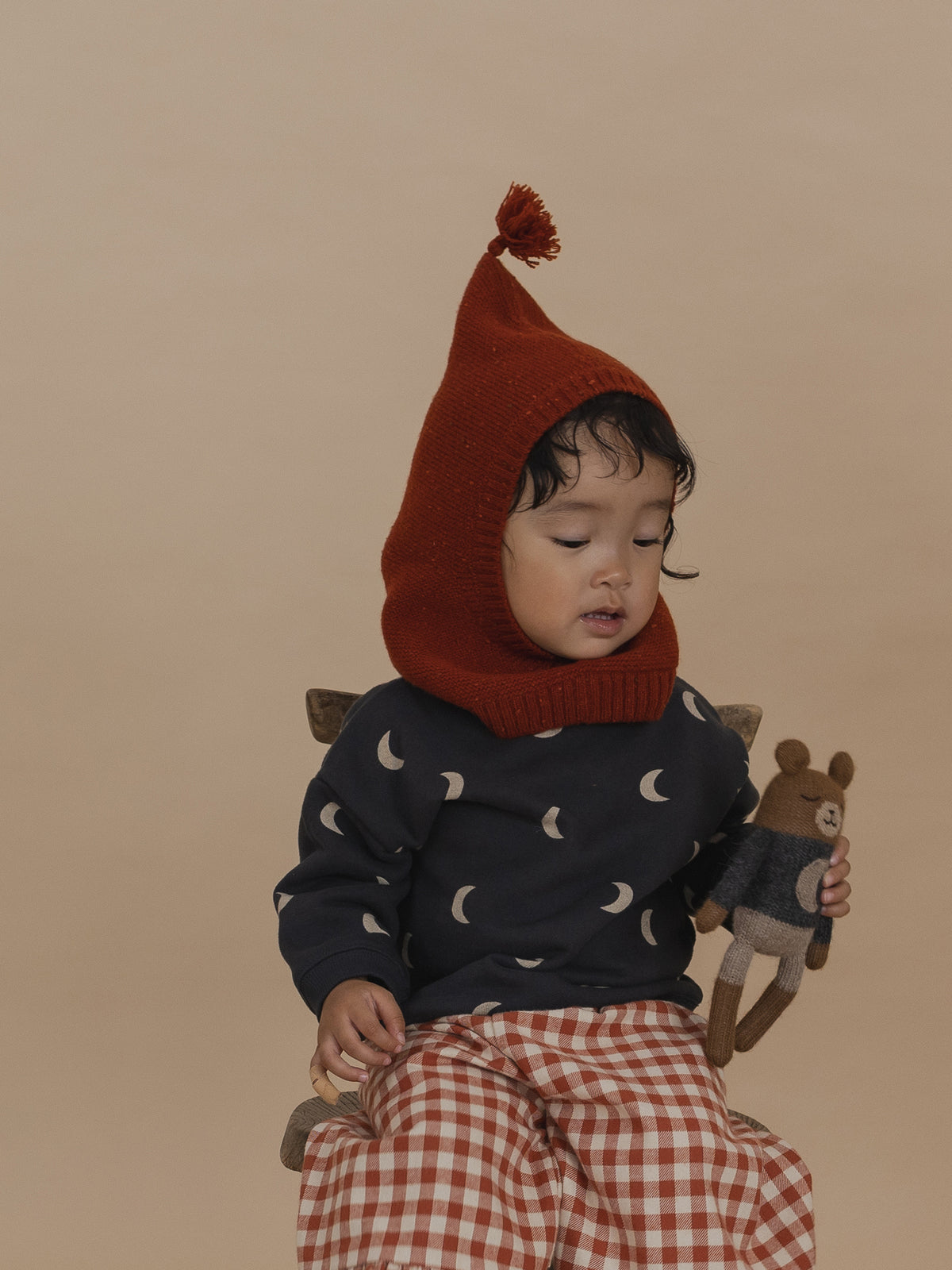 toddler in red wool balaclava with tassel, black moon print sweatshirt, gingham pants, holding toy