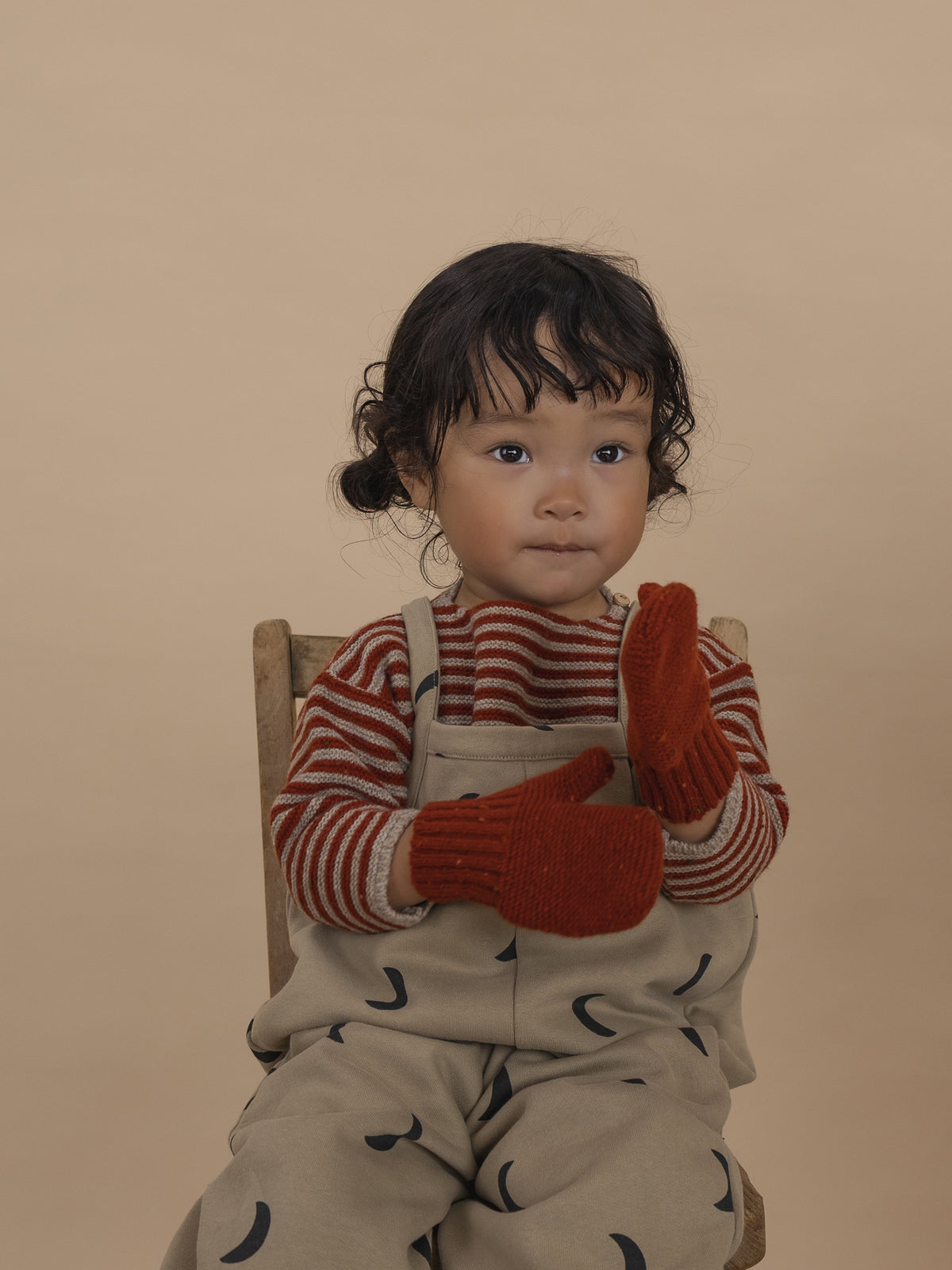 toddler wearing beige dungarees, paprika red wool jumper with matching knitted wool mittens
