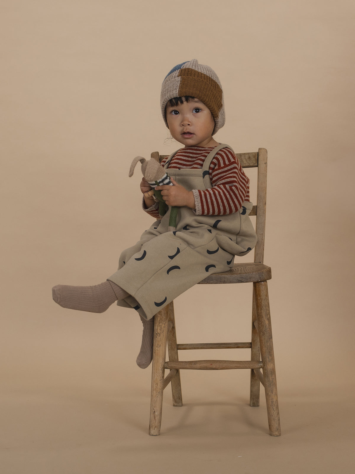 toddler in red striped wool jumper, beige dungarees and beanie, holding plushie and sitting on chair