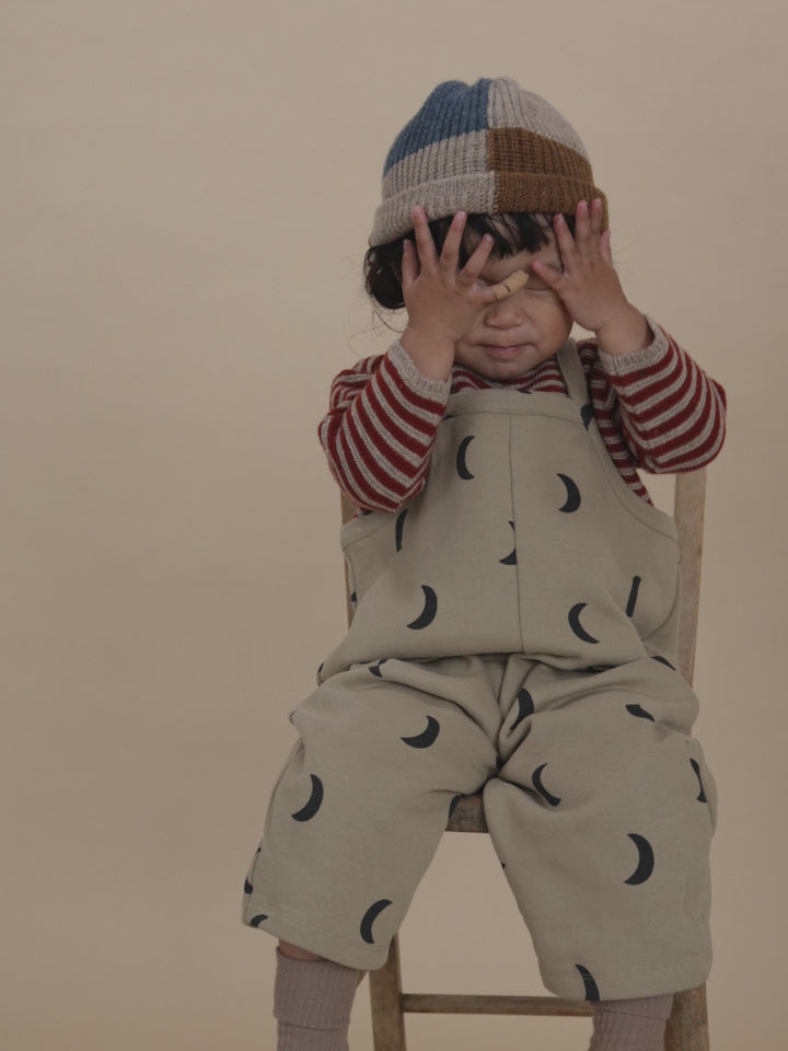 toddler in beige dungarees with black moon print, striped sweatshirt & patchwork beanie, playing peekaboo