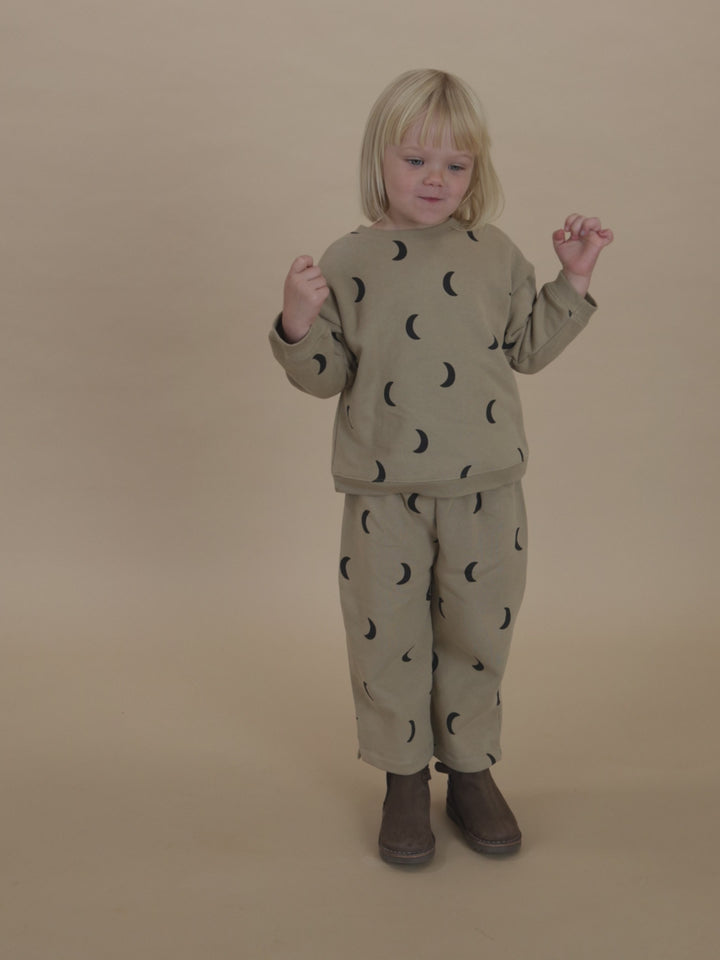 toddler in beige pants with black moon print & matching sweatshirt, posing on one leg with arms out
