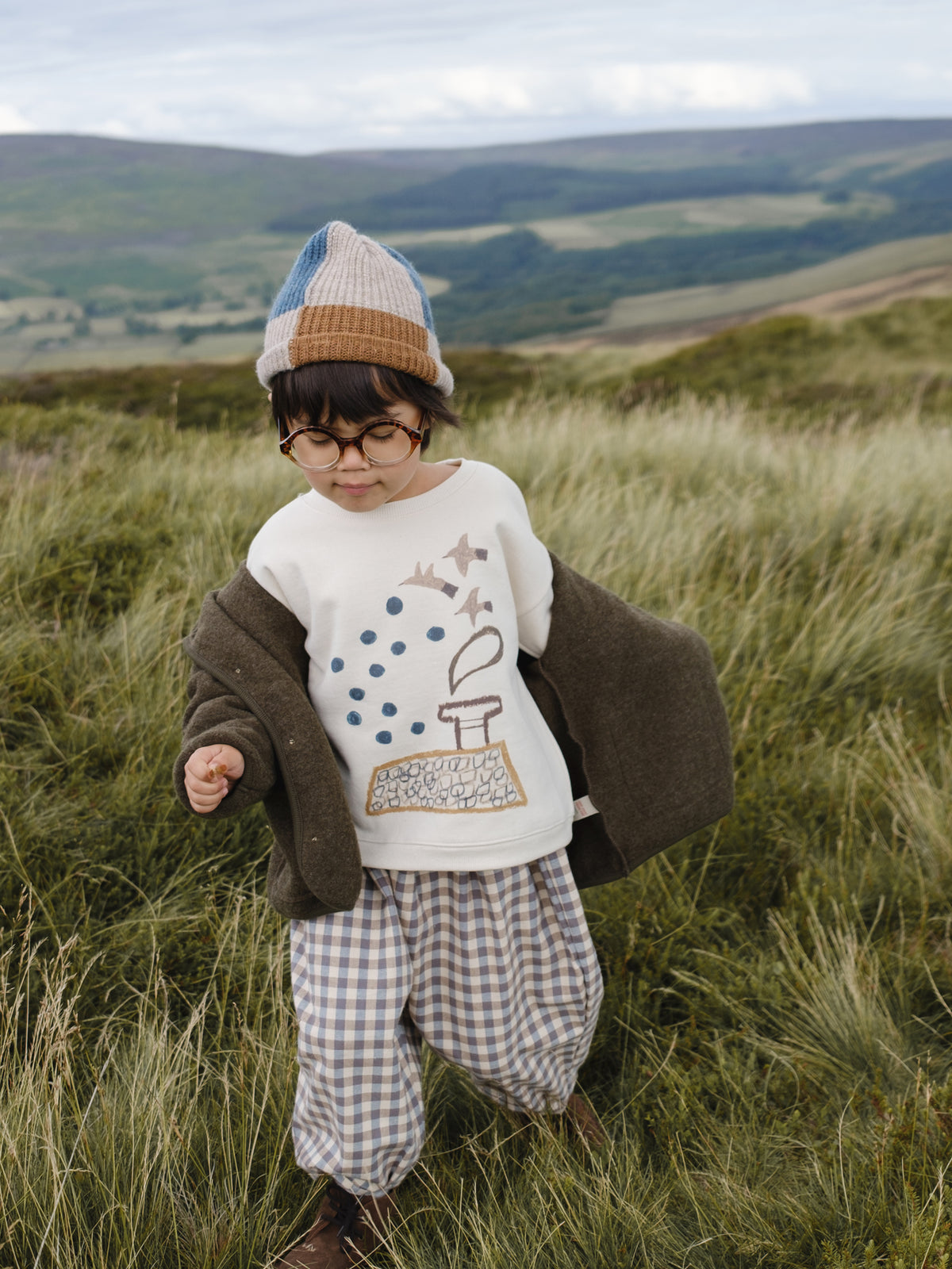 toddler in sweatshirt with bird graphic, gingham pants, patchwork beanie, and wool jacket in grassy meadow