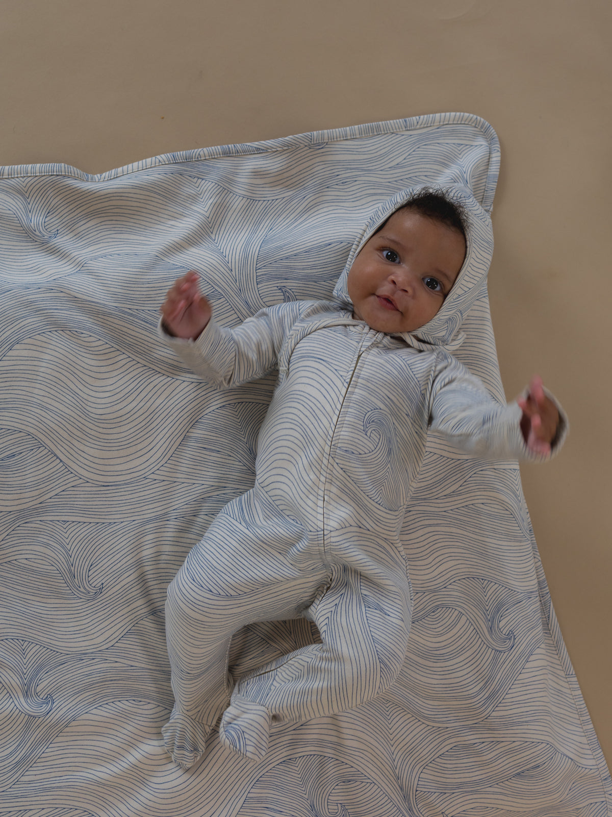 baby in cream pixie bonnet & playsuit with blue line-drawn ocean waves, lying on matching blanket