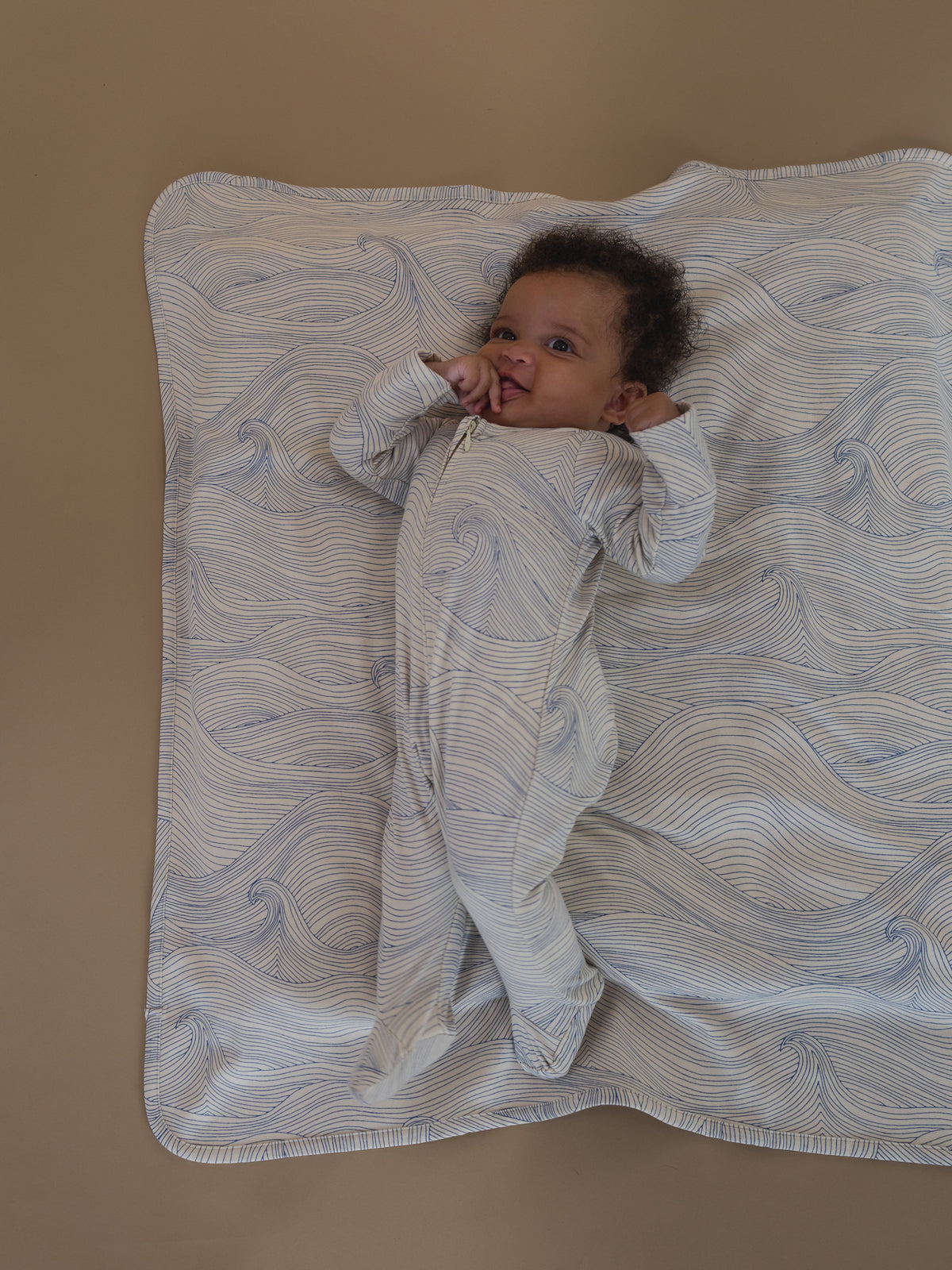 baby in undyed cotton zip sleepsuit with blue line-drawn ocean waves, lying on matching blanket