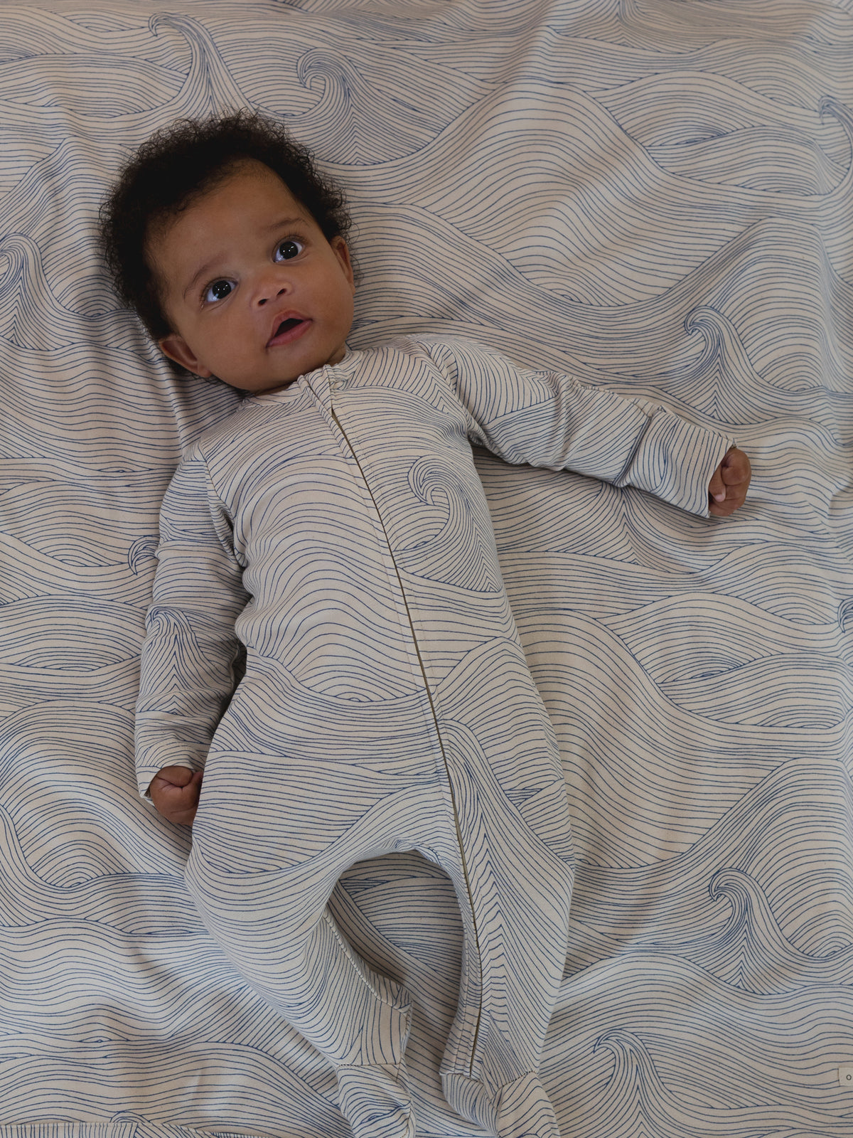 baby in undyed cotton zip sleepsuit with blue Seascape line-drawn ocean waves, and matching blanket