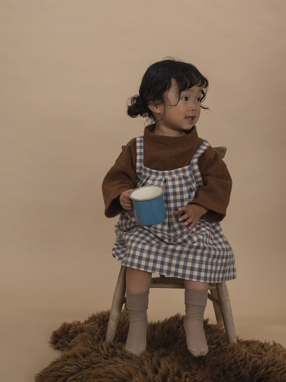 toddler in cream and blue gingham pleated pinafore skirt with brown jumper, sitting on chair with cup