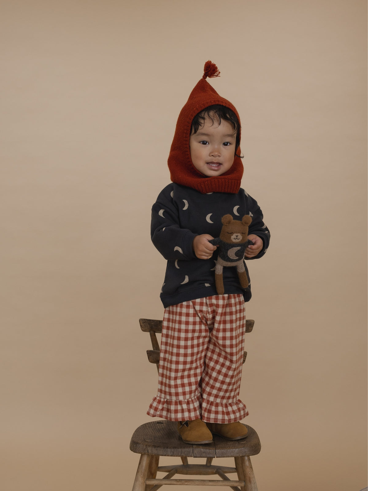 toddler in sweatshirt, gingham pants and balaclava, holding teddy in matching jumper with moon motif