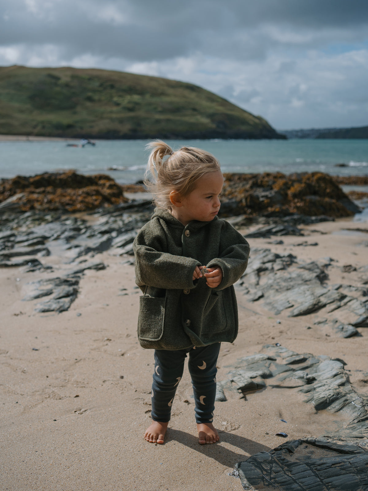 Child in wool jacket and midnight leggings standing on a rocky beach with a body of water and hills in the background
