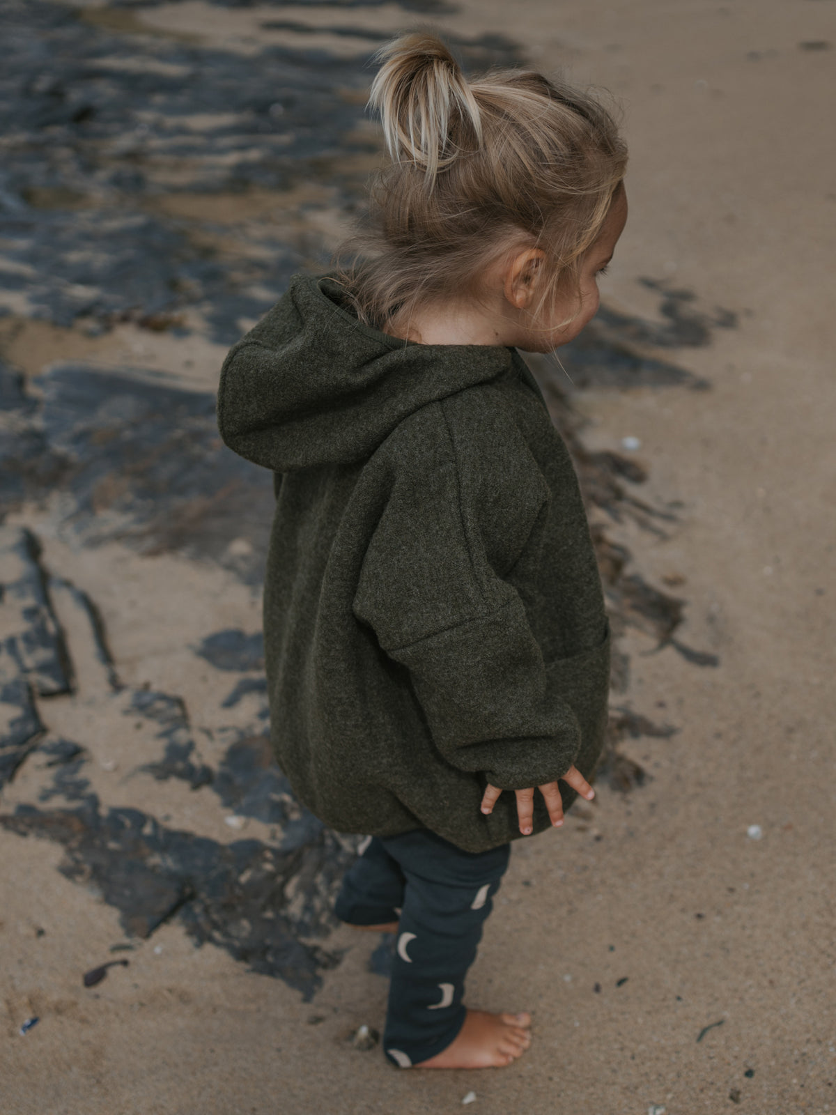 Child wearing a green wool hoodie standing on a sandy beach.