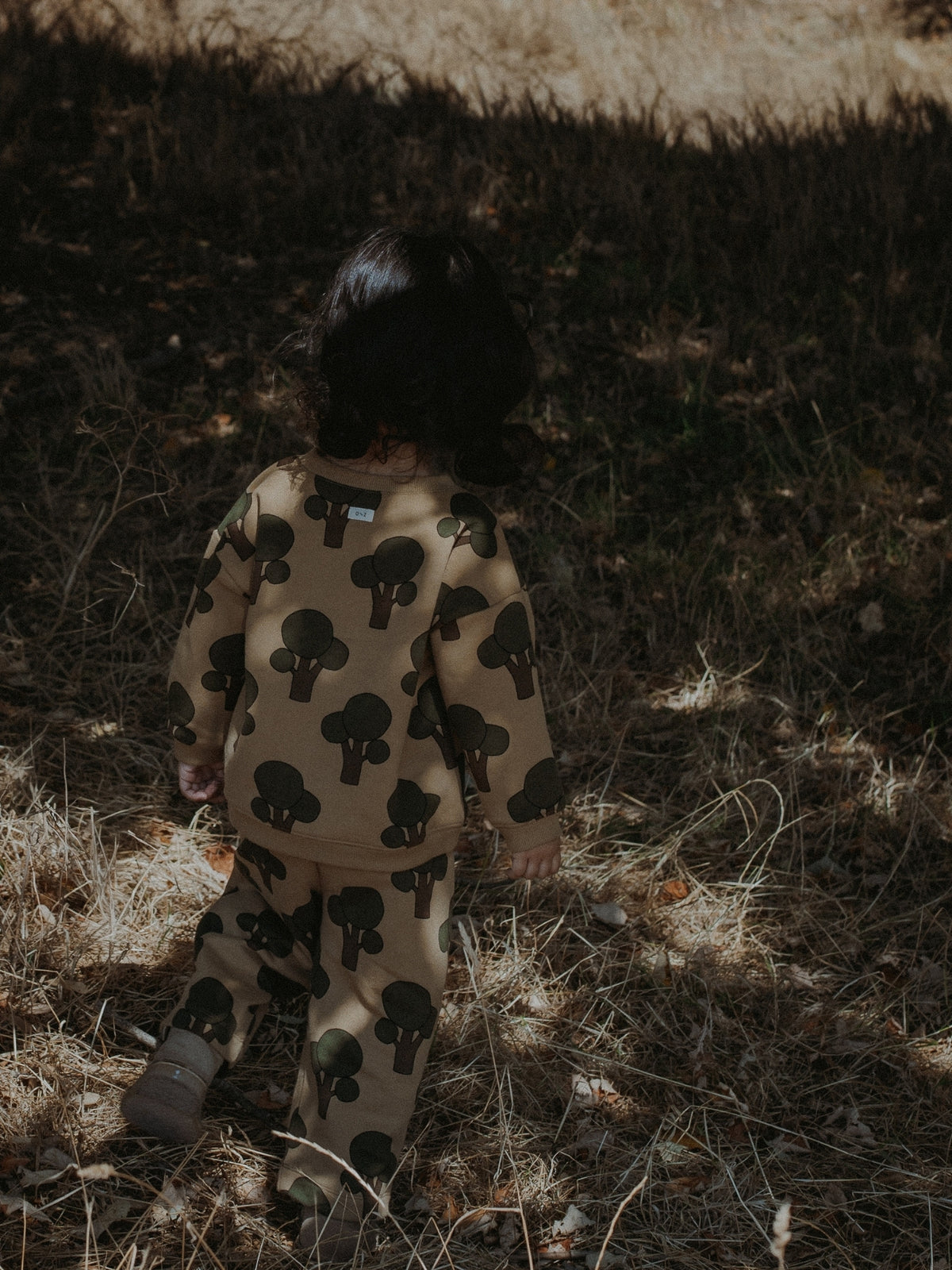 A child seen from behind wearing a tree co-ord set set with a black tree print, standing outdoors on dry grass in dappled sunlight.