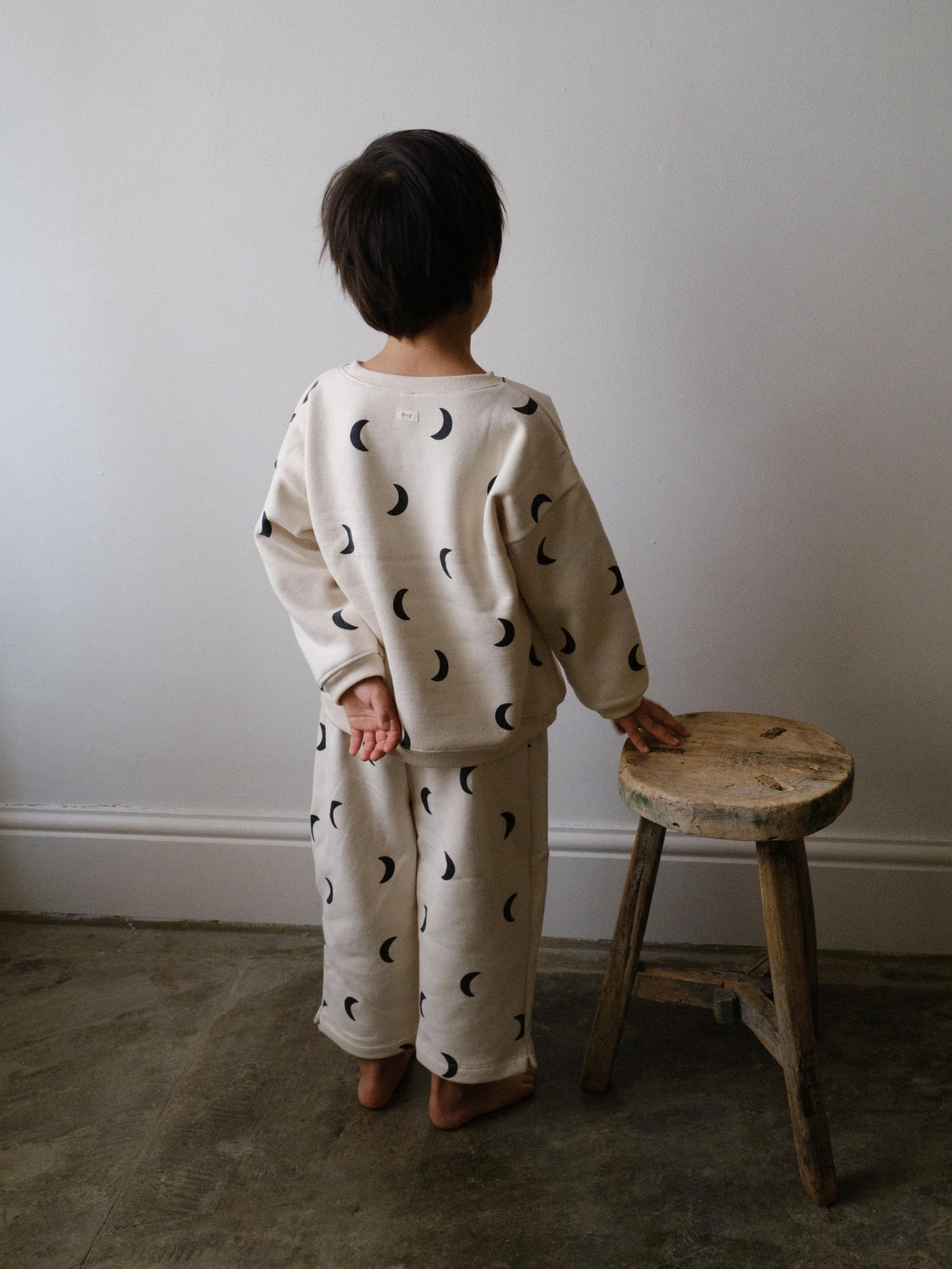 Child wearing a cream-colored outfit with black moon patterns, standing next to a wooden stool against a plain wall.