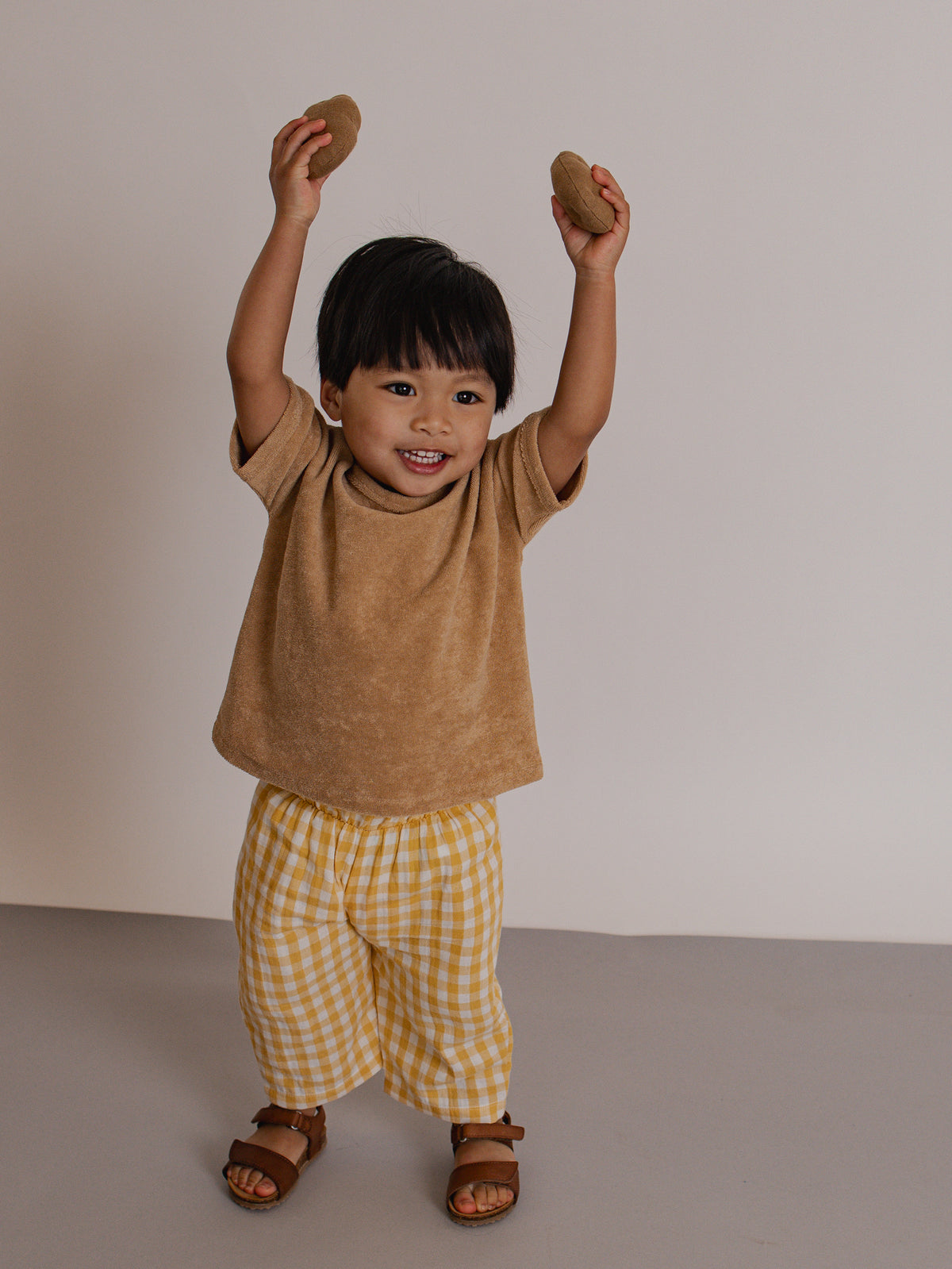 Child wearing a brown shirt and yellow checkered pants holding two stones above their head against a plain background.